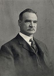 A black-and-white photo of a mustachioed man wearing a dark Victorian-style suit and tie and white shirt