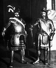 Two men standing in full armor. They have on hats with feathers and are carrying swords