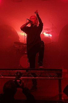 Will Ramos standing onstage, singing into a microphone and raising his other hand, with a red backlight