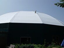 A round, green building with a white tent-like roof stands against a clear summer sky.