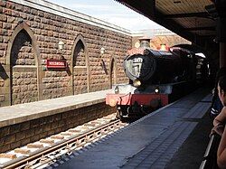 A railway vehicle disguised as a red-colored steam locomotive pulling into an old-fashioned railroad station building.