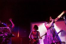 Three women playing instruments on a stage. Left–right: One woman plays a keyboard, the second a guitar, while the third woman sings into a microphone.