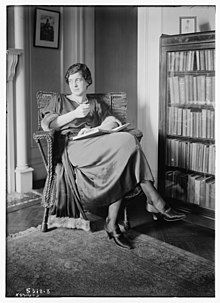 A white woman with dark hair, seated indoors, in front of a bookcase, with a book open in her lap