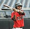 An African-American male wearing a black baseball helmet with a red jersey, displaying two eyes on the front. He is also holding two bats in his hand, one red and the another black.