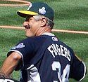 A photograph of a man with a grey moustache facing left and wearing a blue baseball hat with a yellow brim and sticking his tongue out of the side of his mouth