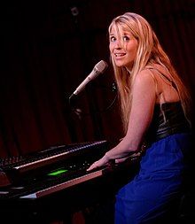 Charlotte Martin wearing a black leather top and dark blue skirt, sitting and playing a keyboard while singing into a microphone, head turned sideways, as if to look at audience.