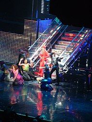 Distant image of a woman. She wears a red suit and a red hat. She is standing in the lowest step of a long metal staircase. She is surrounded by many people who are posing, standing and on the floor.