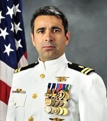 A man in a white naval uniform adorned with medals. Behind him is an American flag.