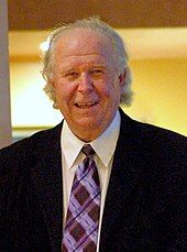 A man with disheveled white hair wearing a black suit jacket and purple tie looks straight ahead and smiles.