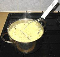 A potato masher in use, with a freshly-prepared pan of mashed potatoes