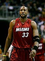 A black bald man wearing a basketball uniform walks on a basketball court toward the camera.