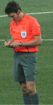 A man wearing a bright orange shirt and grey shorts, writing in a notebook. He has dark hair.