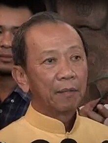 Colour photograph headphones of Sommai Pasi aged 70. he is wearing a smart looking yellow collared shirt. He has dark hair but is slightly balding. the headshot appears to be a cropped image of him in a crown or audience of people