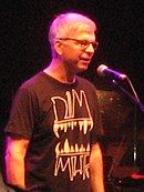 A gray-haired man with glasses and a black shirt standing in front of a microphone