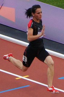 Dana Hussain is seen running on the orange Olympic track. She is in the row second from the outside area of the track. There is a purple border fence surrounding the track. She is wearing a black shirt and shorts, with red details. The number 2 is visible on her shorts from her number sticker, but the other number are out of frame. The number sticker on her shirt is blocked by her arm, as she runs. She is wearing red and white sneakers.