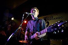 Peter Brewis of the rock band Field Music performing on a stage, playing a guitar and singing into a microphone, as other musicians perform in the darkened background behind him.