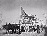 Wagon being pulled by oxen, with large number of people standing in the wagon, one holding a large flag