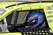 A man in his late 20s wearing a racing helmet and sitting inside a closed-cockpit stock car.