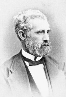 Head and shoulders photograph of a man in a Victorian suit. He has a white beard and large mustache He wears a serious expression and is looking to the left.