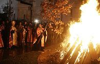 Open-air fire, built with conically arranged long pieces of wood, blazes in the night. Orthodox priest places a long oak sapling with brown leaves on the fire. The priest and the fire are surrounded by a ring of people watching them. In the background, walls of a great church are visible.