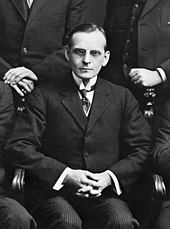 Black and white photo of a middle-aged seated in a chair, wearing a suit and tie