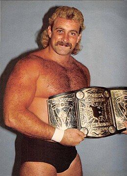 A color photographs of a shirtless man with a mustache holding a mostly-gold professional wrestling championship belt