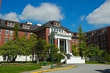 an angle of a large building with leaved trees in the foreground. The building is red in colour with a darkened roof and white coloured entrance.