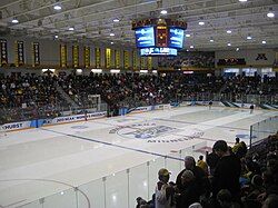 An arena set up for ice hockey. Spectators surround the rink, and the ice is in the middle of being cleaned by Zambonis.