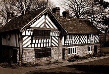 Photograph showing a timber-framed house. There are tow sections to the building at right angles to each other forming a T shape. The ground level of the building has stone walls, whereas the upper floor has wattle and daub infill.