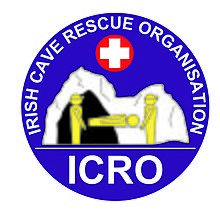 Circular graphic logo, depicting two cavers carrying a casualty on a stretcher away from a cave entrance.