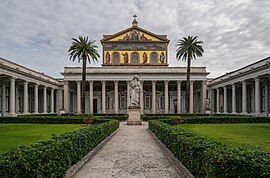 A courtyard with palm trees and a greater-than-lifesized statue of Saint Paul holding a sword in front of the colossal portico of the basilica and a large mural covering the upper facade