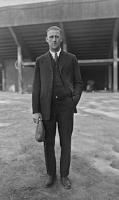 A young man wearing a dark three-piece suit and a dark colored necktie holding a flat cap in his right hand.