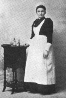 A young woman with dark hair and brown skin, wearing a nurse's uniform, standing next to a small table with bottles on it