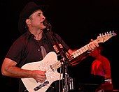 A man wearing a short-sleeved black shirt playing a white guitar and singing into a microphone. The background is black, except for a seated man playing on a keyboard and singing into a microphone.