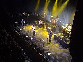 Pop group performing on stage, spotlit, with row of darkened fans in the foreground