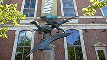 Two Deinonychus figures balance on a claw-like form, in active poses as if they are pursuing prey. In the background is a banner advertising the The Academy of Natural Sciences, and the brick structure and windows of the museum itself. It's late in the afternoon on a sunny day in July, and a sunbeam striking the face of one of the sculpted dinosaurs casts dramatic shadows.