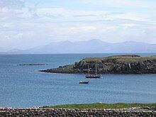 A two-masted yacht and a RIB lie at anchor on a sunny day off a grassy shore, with low cliffs beyond. A skerry lies further offshore to the left with high hills in the distance.