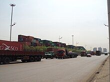 line of heavy trucks going north, background a container yard