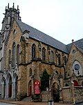 A two-story, brown brick church stands at a street corner, with a tall gray tower looming in the background.