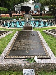 A long, ground-level gravestone reads "Elvis Aaron Presley", followed by the singer's dates, the names of his parents and daughter, and several paragraphs of smaller text. In the background is a small round pool, with a low decorative metal fence and several fountains.