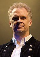 A Caucasian male with silver cropped hair and a goatee looking off camera. He is wearing a white shirt opened at the neck with black jacket.