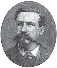 A man with dark hair and a dark, wiry mustache and goatee wearing a white shirt and black jacket