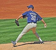 A man wearing a blue cap, blue top, and light-colored trousers, holding a baseball. The back of his shirt reads "Maddox 30"