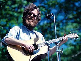 bleached-out image of Loudon Wainwright III wearing sunglasses and playing an acoustic guitar on on outdoor stage