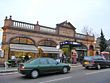 A brown building with a sign that reads "BARONS COURT STATION DISTRICT RAILWAY" under a blue sky with white clouds with two cars in the foreground