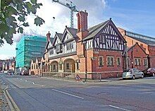 The front wing of the building seen from its corner. It has two storeys; the lower storey is in brick with stone dressings and contains two wide arches in the entrance front; the upper storey is timber-framed and gabled; at both extremities there are tall chimney stacks with spiral decorative brickwork. Behind the entrance wing are larger wings that contain the baths.