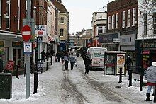 Street scene with pedestrians and vehicles in road lined with shops. There is snow on the ground.