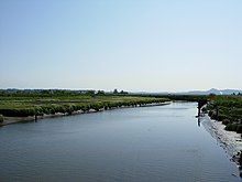 A photograph of a calm river in the middle of rural farmlands, during the day