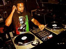 Man sitting in a studio before two turntables and audio turning buttons, adjusting the sound as he listens over earphones.