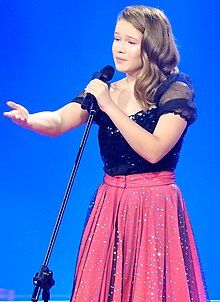 Close-up of Erin Mai in black with a red skirt, holding a microphone and reaching her other arm out